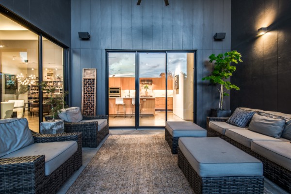 A modern covered patio at dusk with woven seating and warm interior light beyond