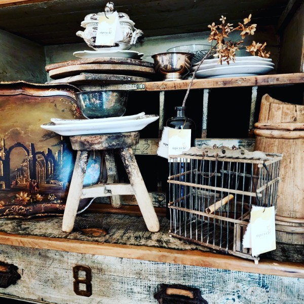 A rustic vignette of a primitive stool, silver tureen and wire basket on a weathered dresser
