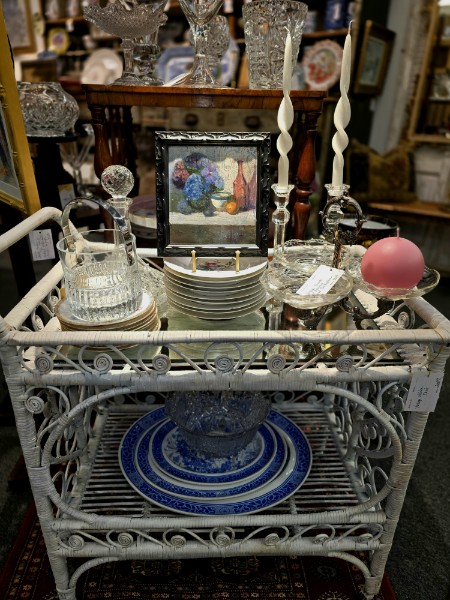 A white wicker bar cart styled with cut crystal, blue and white china and a still life painting